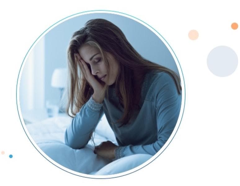 Mujer joven sentada en cama inclnada hacia adelante y con sus manos sobre su estómago debido al dolor estomacal.