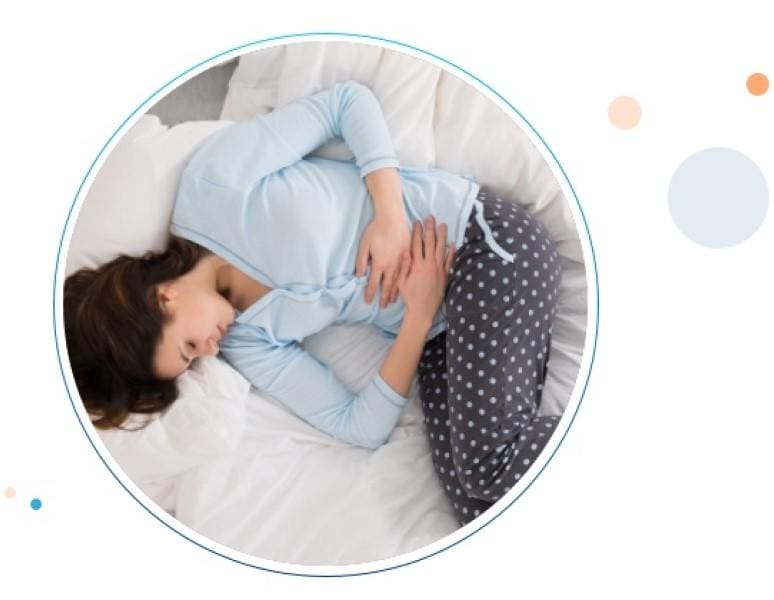 Una mujer joven sufriendo dolor abdominal, recostada en cama con ojos cerrados y las manos en su abdomen.