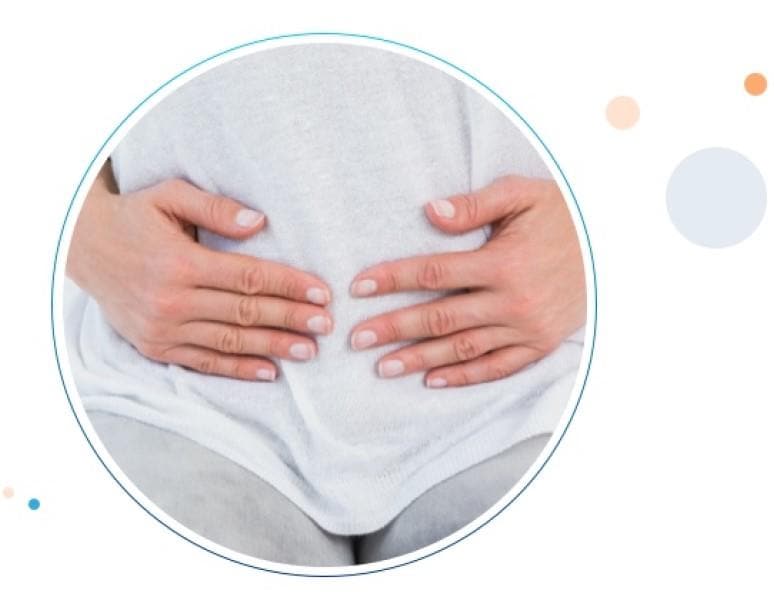 A young woman sitting on her bed with her hands around her belly because of stomach pain. El vientre de una mujer sentada sobre su cama con las manos sobre su abdomen por dolor estomacal.