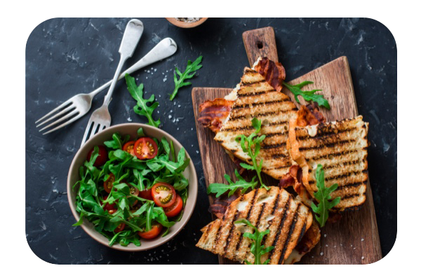 Sandwiches de tocino y queso mozzarela a la parrilla en una tabla de madera junto con un tazón de ensalada – un almuerzo saludable para evitar la indigestión.
